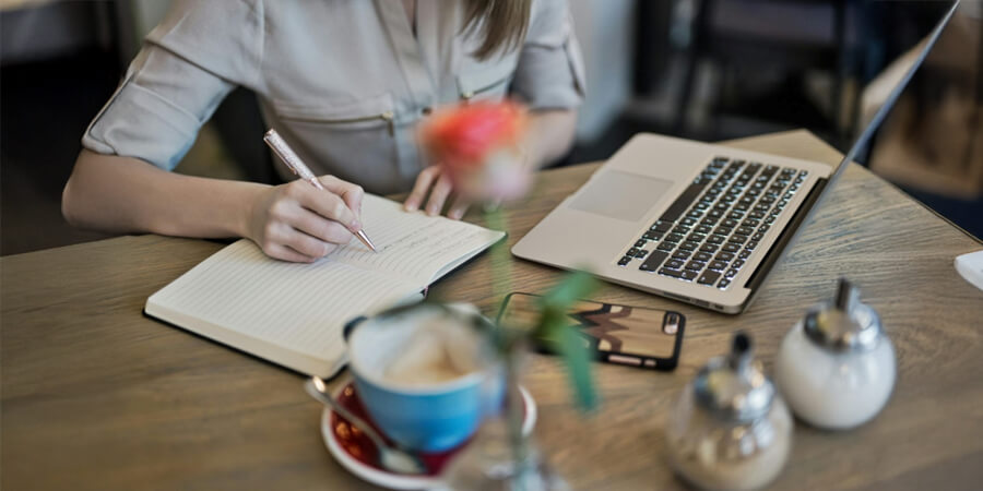 Person writing in a notebook with a coffee, phone and laptop.